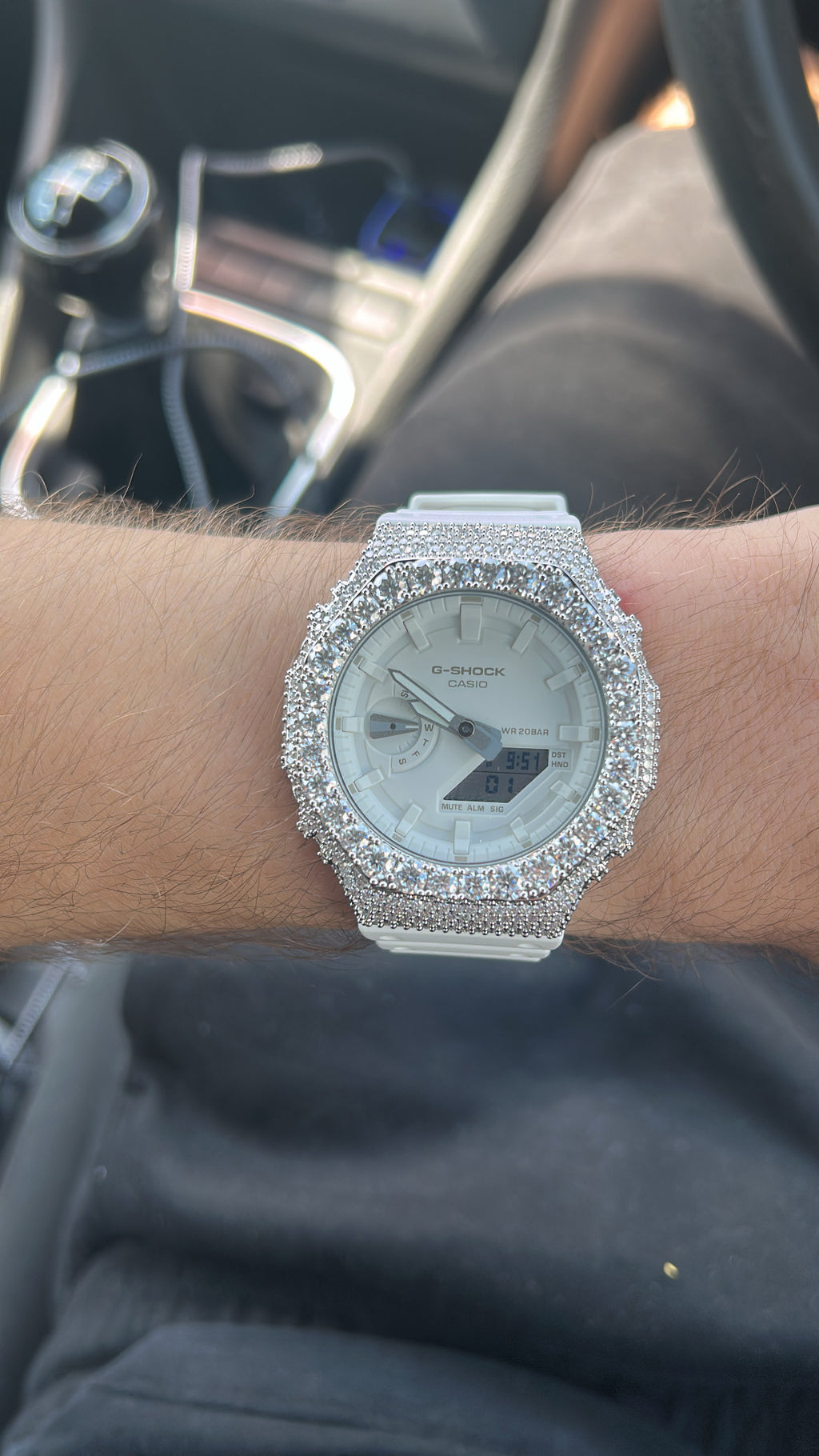 A close-up of a wrist wearing a white Casio watch heavily embellished with sparkling clear stones around the bezel and strap, set against a blurred car interior background.