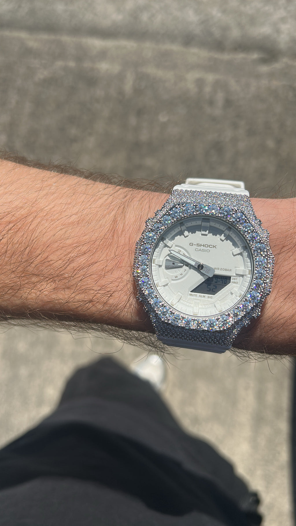 A close-up of a wrist wearing a heavily embellished watch with a silver-toned bezel and case covered in sparkling stones, paired with a white dial and white strap, photographed outdoors against a gray paved background.